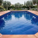 Piscine extérieure avec liner bleu cobalt profond, entourée de pierres en travertin et de végétation méditerranéenne luxuriante